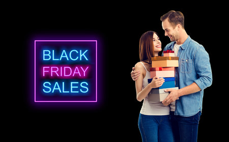Big deals, discounts offers concept - happy young couple holding gift boxes, looking at each other. Black friday sales neon light sign text. Dark background.の写真素材