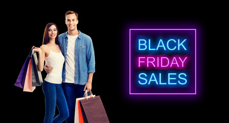 Big deals, discounts offers concept - happy couple with shopping bags. Isolated over dark background. Black friday sales neon light sign text.の写真素材