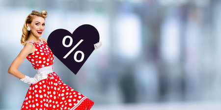 Woman holding paper heart with % sign.  Portrait image of happy pin up girl. Blond model at retro fashion rockabilly sales ad. Valentine or like symbol. Black Friday sale. Blurred mall background.の写真素材