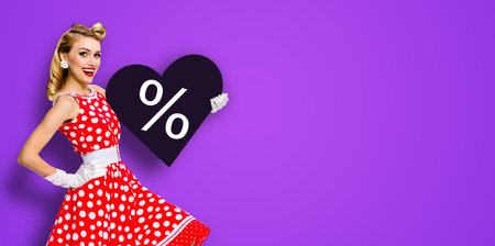 Woman holding paper heart with % sign.  Portrait image of happy pin up girl. Blond model at retro fashion rockabilly sales ad. Valentine or like symbol. Black Friday sale. Violet background.の写真素材