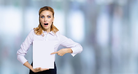 Very happy, excited surprised, astonished business woman with wide opened eyes, mouth showing blank paper signboard. Success ad concept. Copy space empty place for some text. Blurred office backgroundの写真素材