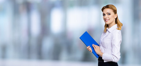 Happy smiling attractive businesswoman, real estate agent copy space for some slogan or text. Confident blond woman holding blue folder, on blurred office background. Blurred office background.の写真素材