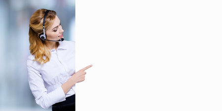 Call Center Service concept - customer support or sales agent. Business woman, caller, adviser phone operator pointing at empty white banner. Help, answering, consulting. Blurred office background.の写真素材