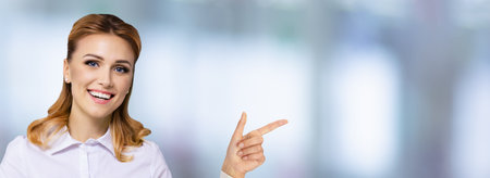 Image of confident woman, showing pointing finger side away. Success in business ad concept. Blurred modern office background. Wide horizontal banner composition with copy space empty area.の写真素材