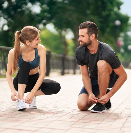Square image of young smiling couple, woman training with man or bearded coach trainer tying shoe laces, before morning jogging, cardio workout outdoors. Fitness, sport, healthy lifestyle concept.の写真素材