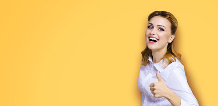 Excited happy smiling businesswoman, showing thumb finger up or like hand sign gesture. Success in business studio concept. Isolated on orange yellow background.の写真素材