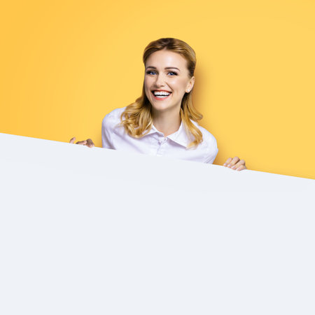 Very happy excited, laughing business woman in white confident clothing standing behind empty banner signboard with copy space area. Success and ad concept. Yellow background. Square image.の写真素材
