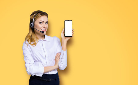 Customer support phone operator in headset holding showing smartphone cell phone mobile with white blank mock up screen, orange yellow background. Consulting and assistance service call centerの写真素材