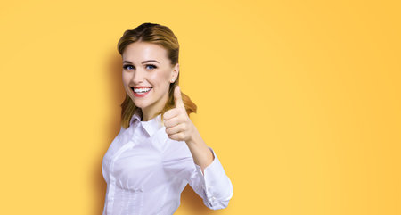 Excited happy smiling businesswoman, showing thumb finger up or like hand gesture. Success in business studio concept. Isolated on orange yellow background.の写真素材