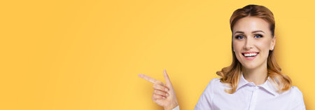 Young attractive confident woman, showing pointing finger at copy space empty text area. Success in business ad concept. Orange yellow background. Wide banner composition.の写真素材