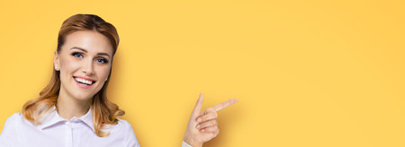 Young attractive confident woman, showing pointing finger at something or copy space empty text area. Success in business ad concept. Orange yellow background. Wide horizontal banner composition.の写真素材