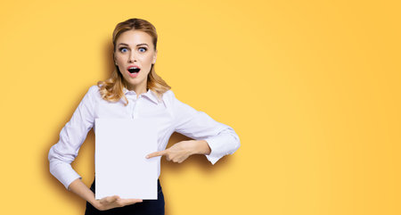 Very happy, excited surprised, astonished businesswoman with wide opened eyes, mouth, showing blank paper signboard. Success in business concept. Copy space place for text. Orange yellow background.の写真素材