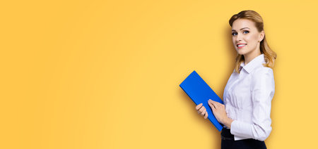 Happy smiling businesswoman, with copy space area for some slogan or text. Confident blond woman in white shirt with blue folder, orange yellow color background. Business concept.の写真素材