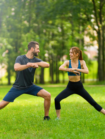 Image of young happy smiling couple, woman training with man or bearded coach trainer, doing squat fit exercise together, look at each other, outdoors. Fitness, sport, workout, healthy lifestyle concept.の写真素材