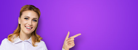 Young attractive confident woman, showing pointing finger at something or copy space empty text area. Success in business ad concept. Violet purple background. Wide horizontal banner composition.の写真素材