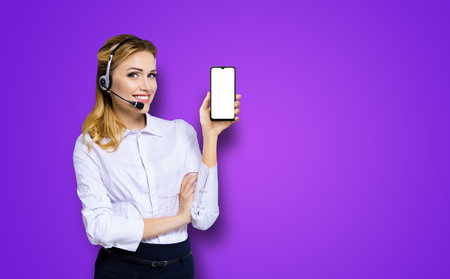 Customer support phone operator in headset holding showing smartphone cell phone mobile with white blank mock up screen, violet purple background. Consulting and assistance service call centerの写真素材