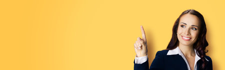 Young brunette woman in black confident suit and violet blouse, showing pointing at copy space for text or imaginary. Business ad concept. Yellow color background. Wide banner image.の写真素材