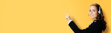 Call Center Service. Customer support or sales agent. Businesswoman or caller or phone operator in black suit showing pointing at copy space. Yellow color background. Help answering, consulting.の写真素材