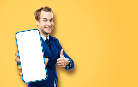 Excited smiling young business man, showing cell phone, mobile smartphone. Happy businessman hold cellphone with white screen, isolated on orange yellow color background. Thumb up. Great app ad concept.の写真素材