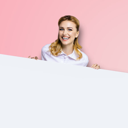 Very happy excited, laughing business woman in white confident clothing standing behind empty banner signboard with copy space area. Success and ad concept. Pink color background. Square.の写真素材