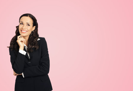 Happy smiling looking up thinking, thoughful woman in black confident suit, copy space area. Business studio concept. Rose pink color background. Attractive brunette businesswoman.の写真素材