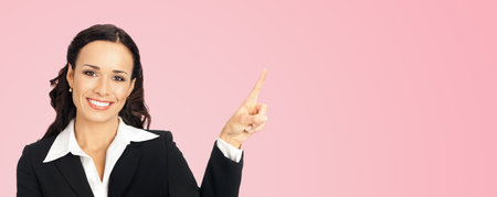 Young happy smiling woman in black confident suit, showing pointing at copy space. Business concept. Rose pink color background. Brunette businesswoman. Wide.の写真素材