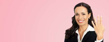 Businesswoman in black confident suit showing four fingers, over rose pink color background. Happy smiling gesturing brunette woman at studio. Business success concept.の写真素材