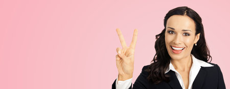 Businesswoman in black confident suit showing two fingers or victory gesture, over rose pink color background. Happy smiling gesturing brunette woman at studio. Business concept.の写真素材