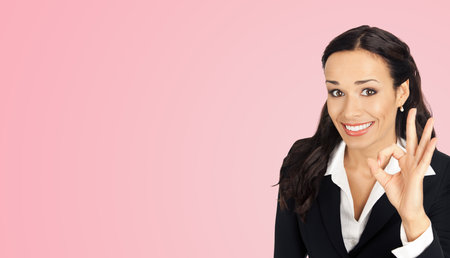 Portrait of businesswoman in black confident suit showing ok okay gesture, over rose pink colour background. Happy smiling brunette woman at studio. Business success concept.の写真素材