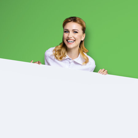 Very happy excited, laughing business woman in white confident clothing standing behind empty area banner signboard. Success and ad concept. Bright green background. Square image.の写真素材