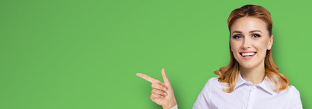 Young attractive confident woman, showing pointing finger at copy space empty text area. Success in business ad concept. Bright green background. Wide banner composition.の写真素材