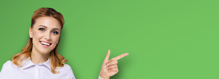 Young attractive confident woman, showing pointing finger at something or copy space empty text area. Success in business ad concept. Bright green background. Wide horizontal banner composition.の写真素材