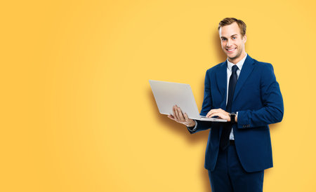 Happy smiling businessman in blue confident suit, working with laptop, over vivid yellow colour background. Handsome young man at studio concept picture. Copy space for ad text.の写真素材