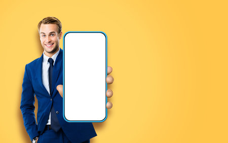 Excited smiling young business man, showing cell phone, mobile smartphone. Happy businessman hold cellphone with white screen, isolated over orange yellow color background. App advertisement concept.の写真素材