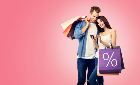 Holiday sales actions, rebates, discounts offers concept image - happy couple with shopping bags, looking at mobile smart phone, isolated on vivid rose pink background. % signの写真素材