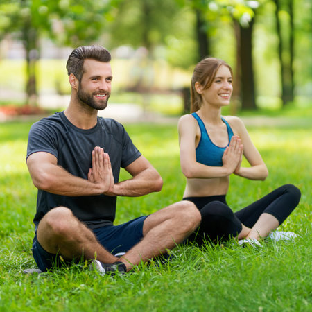 Full body image - young smiling couple  sitting in lotus position or woman practicing with man, male bearded coach trainer, doing yoga exercises or meditating together, after outside training. Wide composition.の写真素材