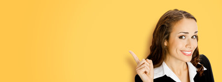 Cheerfully smiling executive woman in black confident suit, pointing showing indicating. Business concept. Yellow color background. Brunette businesswoman. Wide image.の写真素材