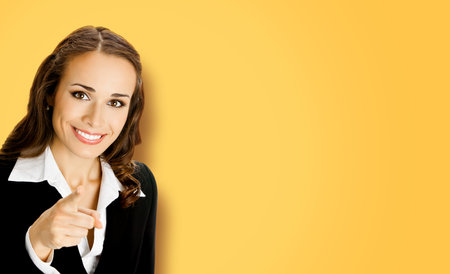 What about You? Cheerfully smiling woman in black confident suit pointing finger at viewer, copy space for text. Business concept. Yellow color background. Brunette businesswoman. Wide.の写真素材