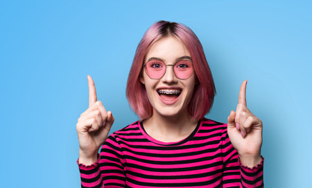 Excited pink woman in braces, opened mouth, wear sunglasses glasses spectacles advertise peeking showing area for sales slogan text, isolated blue background. Dental care, ophthalmology ad concept.の写真素材