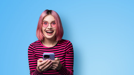 Excited surprised shocked astonished very happy pink funny woman wear braces sunglasses eye glass spectacles open mouth hold typing cell phone cellular smartphone cellphone isolated blue background.の写真素材