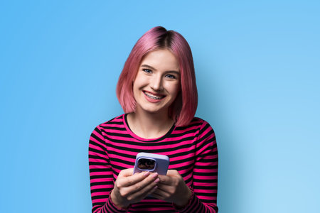 Excited surprised shocked astonished happy pink hair funny woman wear braces opened mouth holding typing cell phone cellular smartphone cellphone, isolated blue plain studio wall background.の写真素材