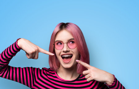 Dental dent care ad concept image - look up pink funny beautiful young woman in metal braces wear sunglasses, pointing white teeth smile. Isolated blue wall background. Positive optimistic.の写真素材