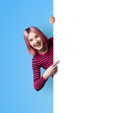 Excited pink woman in braces, opened mouth stand behind, peep from blank empty banner mockup sign board signboard, advertise show area for sales slogan text, isolated blue background. Dental care adの写真素材