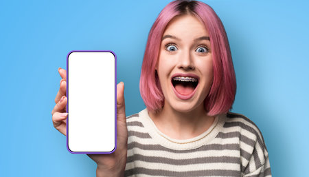 Shock astonished very happy pink woman wear braces, opened mouth, show cell phone, modern smartphone mobile phone cellphone with blank white mock screen, isolated blue wall background. Online offer ad.の写真素材