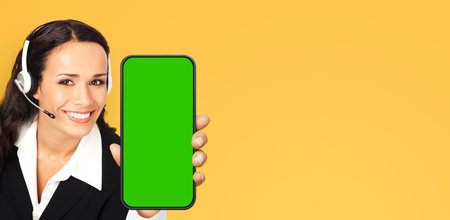 Call Center Service. Customer support, sales phone agent. Businesswoman in headset show cellphone mobile green chroma key mockup screen. Business woman hold smartphone, isolated yellow background.の写真素材