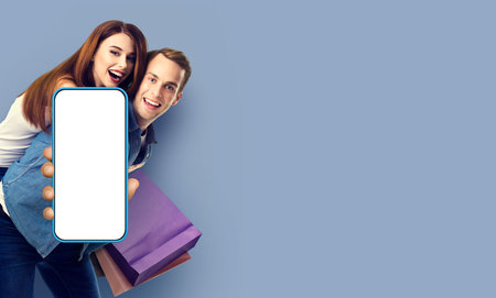 Holiday sales actions, rebates, discounts store retail, cash back offer, consumer concept - happy excited young couple hold shopping bags and cell mobile smart phone with mock up white screen, on greyの写真素材