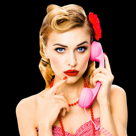 Portrait studio image - pensive, puzzled or thinking beautiful blond woman holding phone tube. Unhappy pin up girl. Retro vintage ad concept. Isolated against black background. Square composition.の写真素材