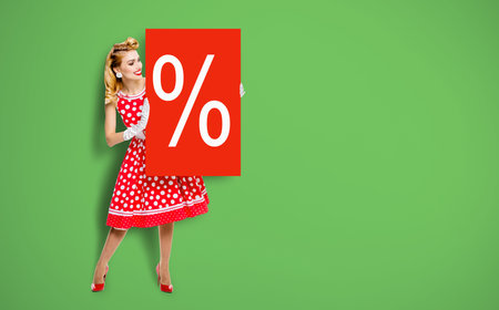 Beautiful woman wear pinup dress hold, show red board percents % sign. Full body pin up girl with advertise offer, isolated against green background. Sales, discounts, rebates ad concept.の写真素材