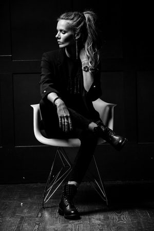 Sexy young model in dark clothes, sitting on a white chair and posing on a dark blue background. Watch on hand. Fashion, Beauty, Styleの写真素材