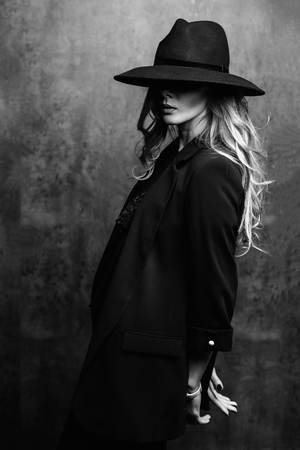 Beautiful young mysterious blond girl in black hat and black jacket on gray background. Eyes are covered with a hat. Manicure - long red nails, nail polish. Fashion, beauty.の写真素材